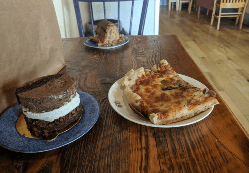 A slice of pizza on a plate next to a layered chocolate dessert on a blue plate, set on a wooden table.