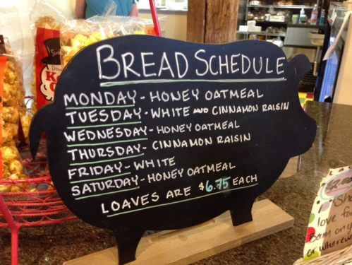 Chalkboard menu listing a weekly bread schedule with flavors and prices, shaped like a pig.