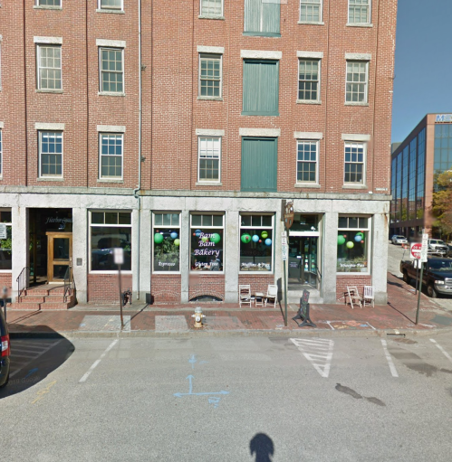 A brick building with large windows displaying a bakery, green balloons, and outdoor seating in a city street.
