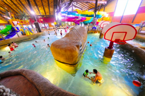 Indoor water park scene with slides, a rock feature, and families enjoying the pool. Brightly colored and lively atmosphere.