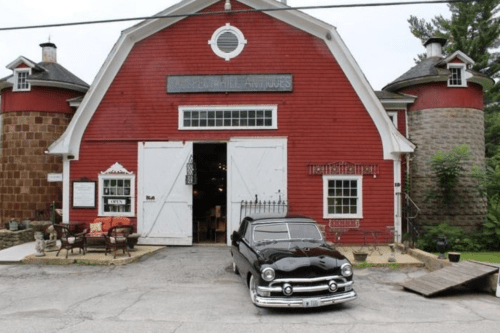 A vintage black car parked in front of a red barn with white doors and a sign reading "Hill Animals."