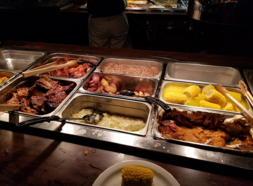 A buffet spread featuring various dishes, including meats, vegetables, and corn, with serving utensils in metal trays.