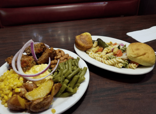 A plate of fried chicken, green beans, corn, potatoes, and macaroni, alongside a pasta salad and a bun.