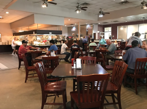 A busy diner with wooden tables and chairs, patrons eating and chatting, and a visible kitchen area in the background.