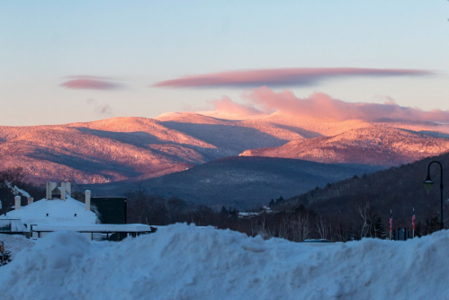 Snow-covered mountains bathed in soft pink light during sunrise, with a clear sky and fluffy clouds above.