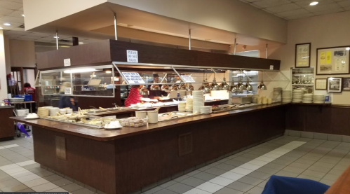 A buffet restaurant with a serving area, plates stacked, and staff preparing food behind a glass partition.