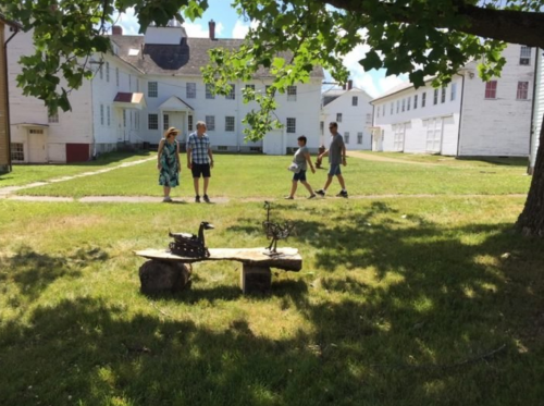 A group of four people walks on a grassy area with buildings in the background and a wooden sculpture in the foreground.