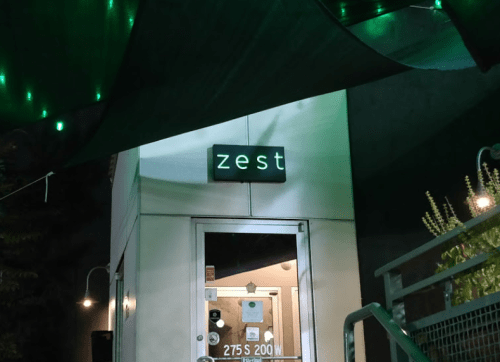 A dimly lit entrance with a sign reading "zest" above the door, surrounded by green lights and a canopy.