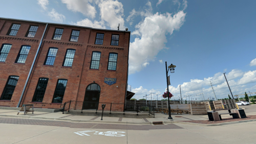 A red brick building with large windows, outdoor seating, and a blue sky with clouds in the background.