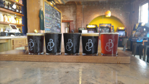 A flight of five craft beers in branded glasses on a wooden tray, set in a cozy bar with arcade games in the background.