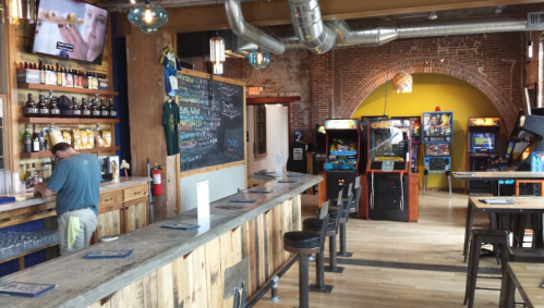 A rustic bar with a wooden counter, arcade games, and a chalkboard menu in a cozy, well-lit space.