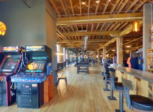A spacious indoor venue featuring arcade games, a pool table, and a bar with wooden floors and exposed beams.