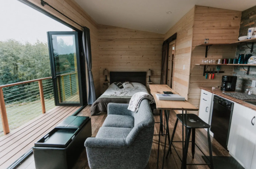 Cozy wooden interior of a modern cabin with a bed, seating area, and kitchenette, featuring large windows and a balcony.