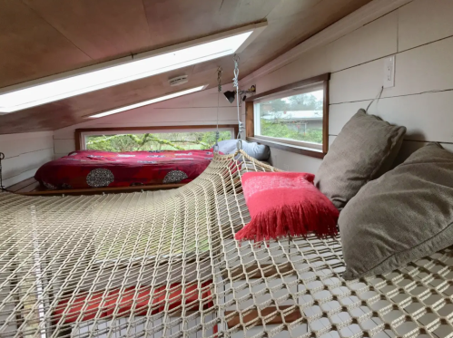 Cozy loft space with a hammock, pillows, and a bed, featuring skylights and a view of greenery outside.
