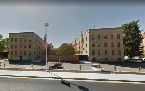 A brick building with multiple windows, surrounded by a parking lot and trees, under a clear blue sky.