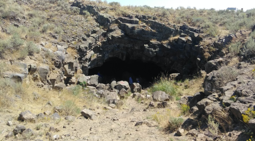 A rocky cave entrance surrounded by dry grass and shrubs, with people visible inside the cave.