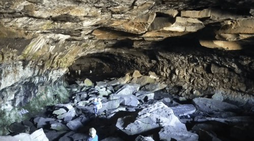 A dark cave interior with rocky terrain and two small figures exploring among large stones.