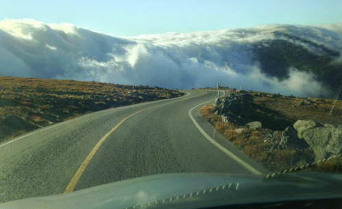 A winding road leads through a landscape with clouds rolling over hills, creating a dramatic, scenic view.