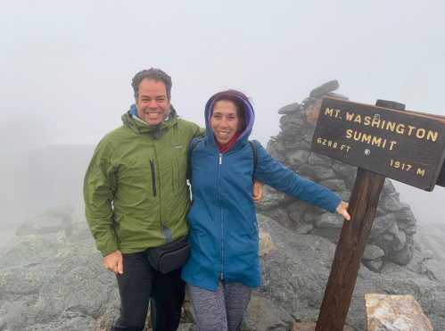 Two people stand at the summit of Mt. Washington, smiling in front of a summit sign, with foggy surroundings.