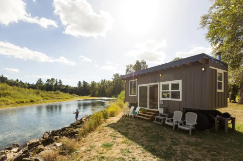 A small modern cabin by a river, with a person fishing nearby under a sunny sky and scattered clouds.