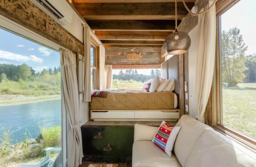 Cozy interior of a tiny home with a bed, sofa, and large windows overlooking a serene landscape by the water.