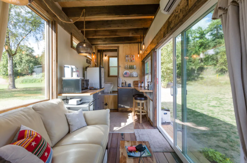 Cozy interior of a tiny house with a sofa, kitchen area, and large windows overlooking a green outdoor space.