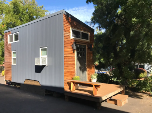 A small modern tiny house with a wooden deck, large windows, and a mix of metal and wood siding, surrounded by greenery.