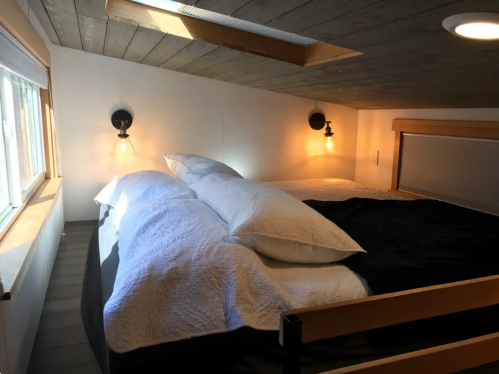 Cozy loft bedroom with a large bed, soft bedding, and warm lighting from wall sconces. Skylight above adds natural light.