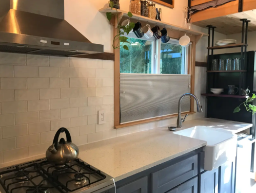 A modern kitchen with a gas stove, farmhouse sink, and large window, featuring plants and dishware on shelves.