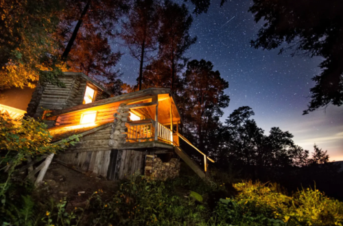 A cozy log cabin illuminated at night, surrounded by trees under a starry sky.