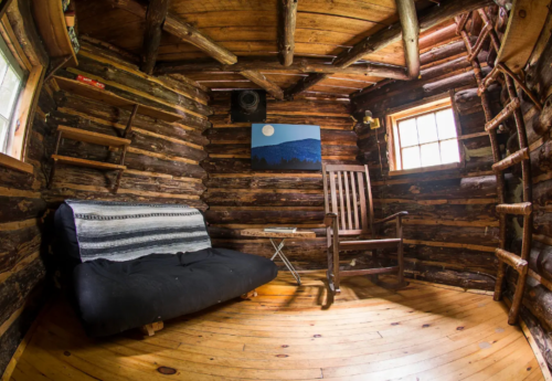Cozy wooden cabin interior with a futon, rocking chair, and a scenic painting on the wall. Warm, rustic atmosphere.