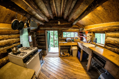 Cozy rustic kitchen with wooden beams, a stove, sink, and a view of nature through an open door.