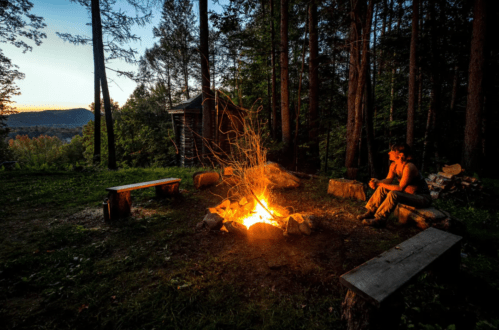 A person sits by a campfire in a forest, surrounded by trees and benches, as the sun sets in the background.