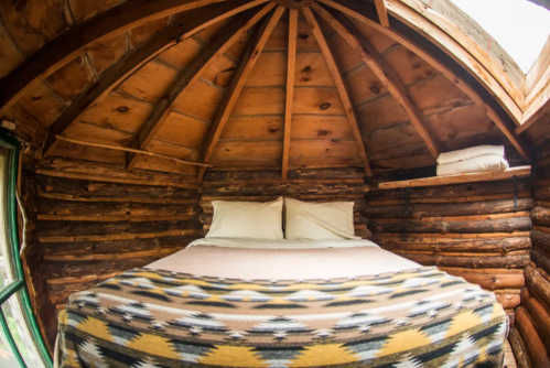 Cozy interior of a log cabin with a bed, wooden walls, and a patterned blanket. Natural light filters in.