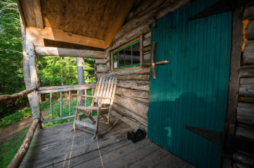 A rustic wooden cabin with a green door and a wooden chair on the porch, surrounded by trees.