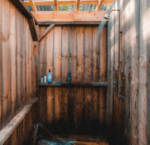 An outdoor shower with wooden walls, featuring a showerhead and shelves with various toiletries.