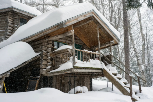 A cozy log cabin covered in snow, nestled among trees in a winter landscape. Snowflakes are gently falling.