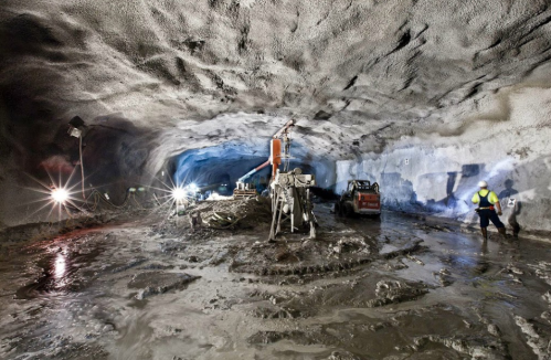 A large underground tunnel with machinery, workers, and wet ground, illuminated by bright lights.