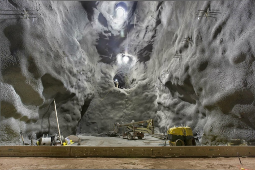 A cavernous underground space with rocky walls, construction equipment, and a worker suspended above.
