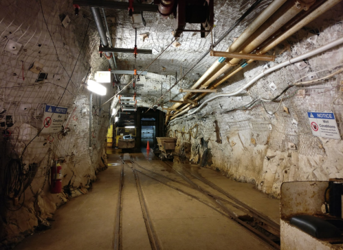 A dimly lit underground tunnel with metal tracks, safety signs, and a mining cart in the background.