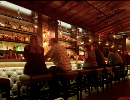 A dimly lit bar scene with patrons sitting on stools, surrounded by shelves of colorful bottles.