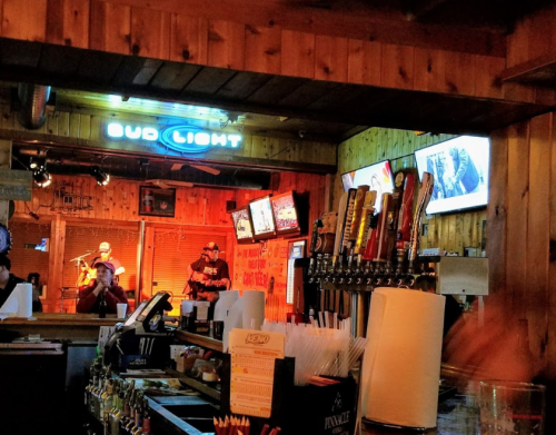 A lively bar scene with a musician performing, illuminated by neon lights and surrounded by beer taps and screens.