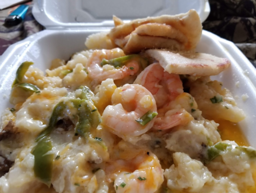 A close-up of a takeout container filled with shrimp, green peppers, and a creamy sauce over a bed of mashed potatoes.