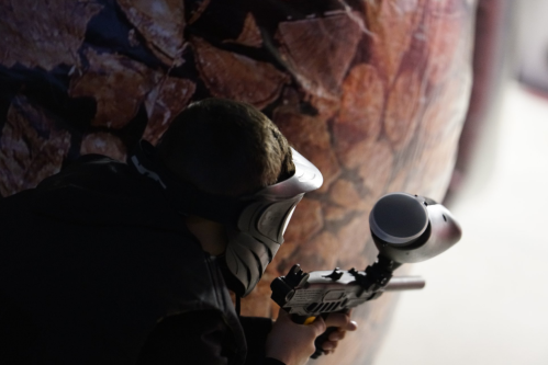 A person in a mask aims a paintball gun behind a large, textured barrier.