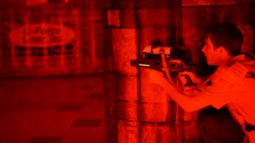 A person crouches behind a pillar, aiming a laser tag gun in a dimly lit, red-themed arena.