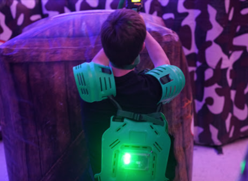 A child wearing a green laser tag vest and arm guards aims a laser gun in a colorful, themed arena.
