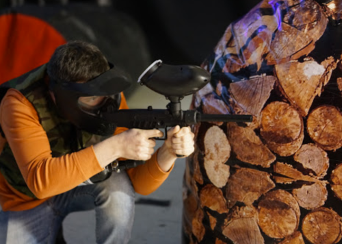 A person in a mask aims a paintball gun at a wooden log target in an indoor paintball arena.