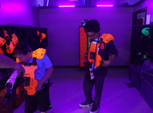 Two kids in a dark room with neon lights, wearing orange laser tag gear and preparing for a game.