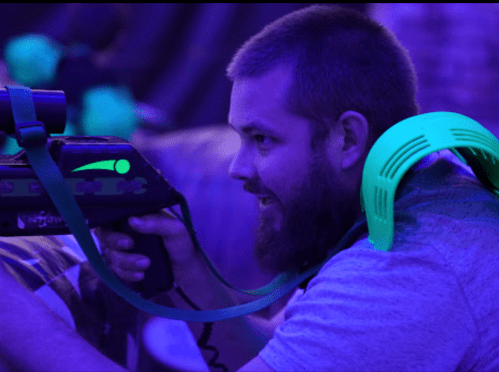 A man with a beard aims a laser tag gun, wearing a green harness, in a dimly lit, colorful environment.