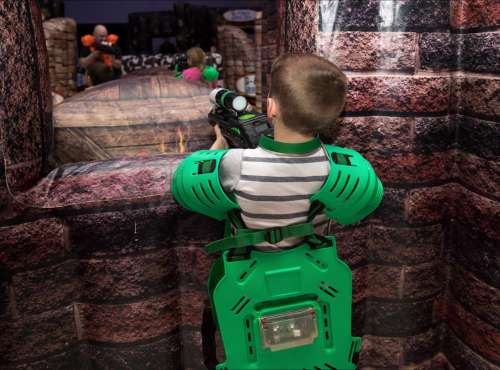 A child in green protective gear aims a toy gun through a window in a play area designed for laser tag.